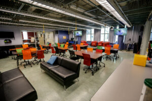 An empty, vibrant collaborative space, possibly a learning lab or flexible classroom, featuring a mix of colorful tables and chairs, comfortable sofas, and lounge seating. Large display screens are mounted on the walls, and decorative fairy lights are strung across the industrial-style ceiling.