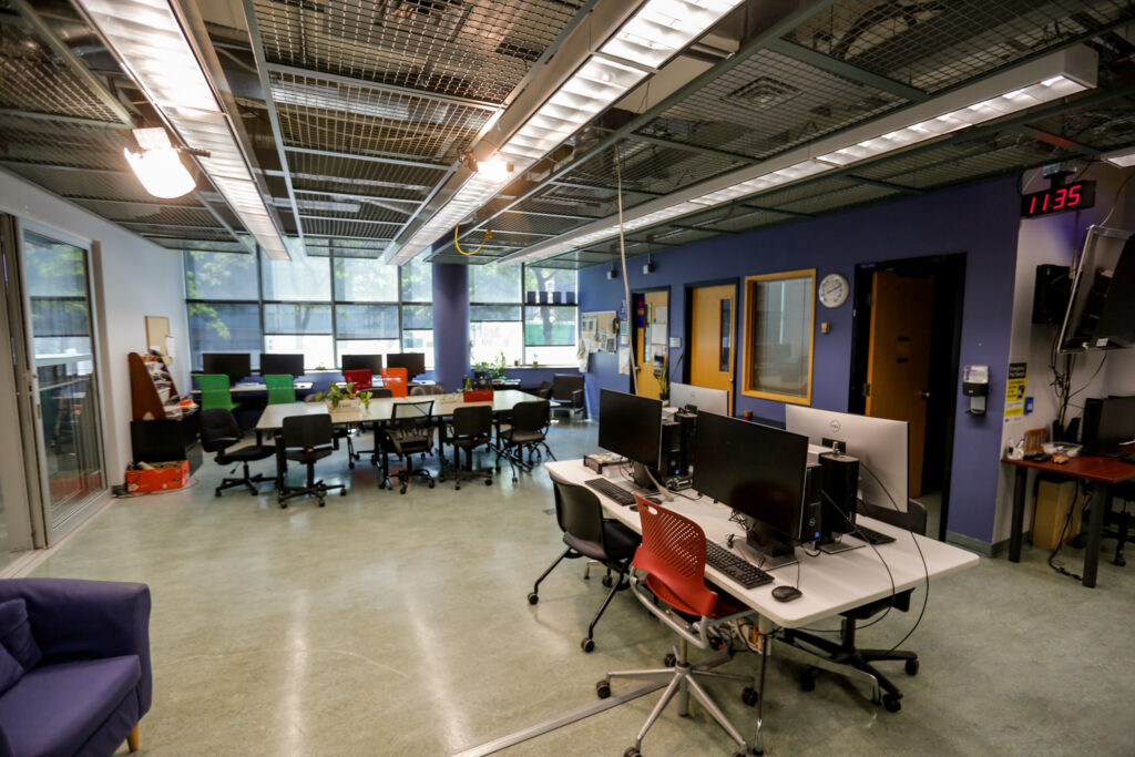 A bright, modern collaborative workspace featuring multiple computer stations, a large central table for group work, and a lounge area with a purple sofa. The room has large windows, blue walls, and an industrial ceiling.