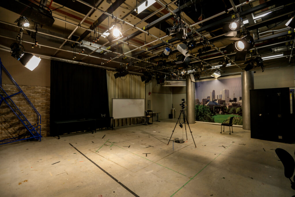 An unoccupied professional TV studio setup, featuring a grid of bright studio lights, a camera on a tripod, and a concrete floor marked with stage tape. Backdrops include a black curtain, a whiteboard area, and a large city skyline mural.