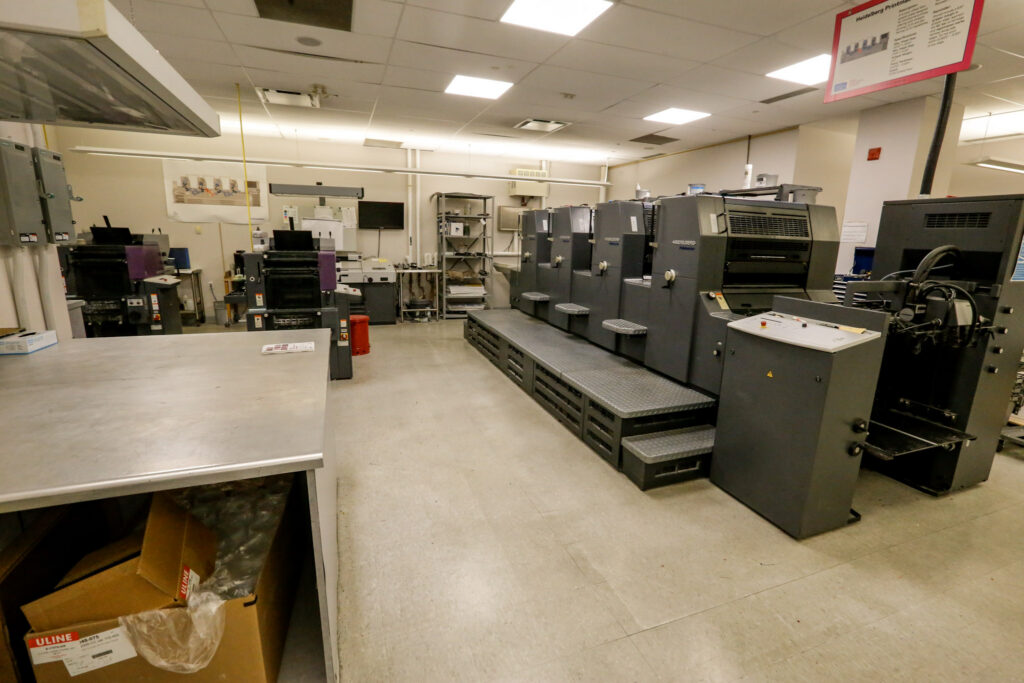 An interior view of a commercial print shop showing a multi-unit Heidelberg printing press on a raised platform, with other graphic arts equipment and work areas.