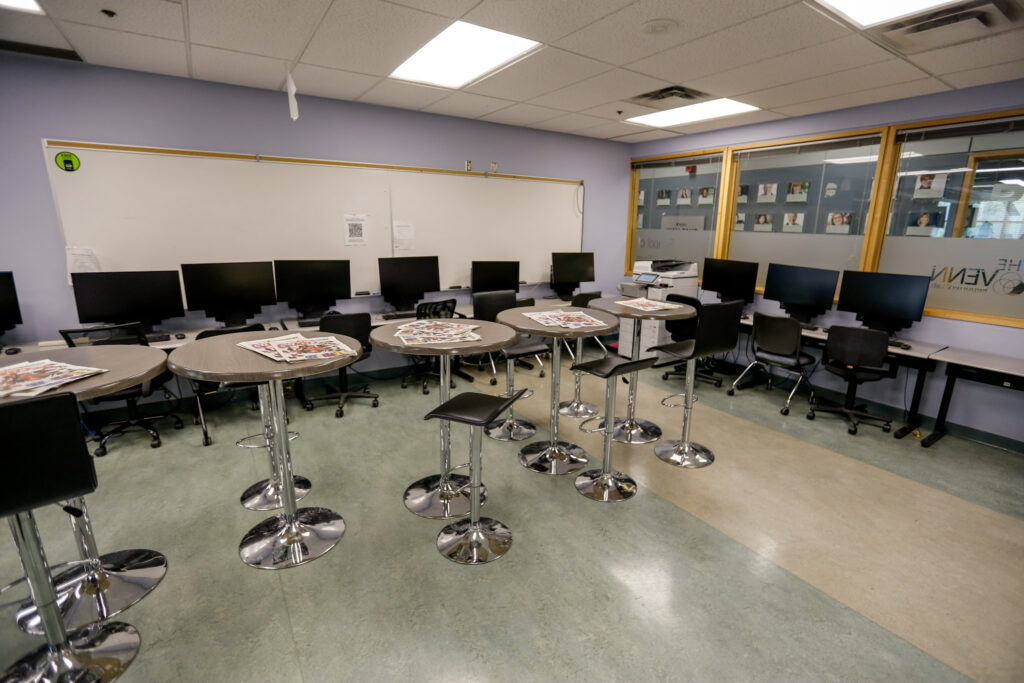 A modern, empty classroom or computer lab featuring tall round tables with magazines and rows of computer workstations along the walls. Large windows look into an adjacent space.
