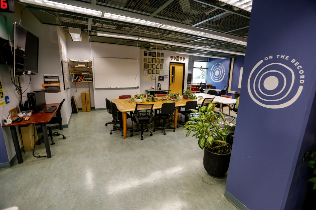 A modern media lab or newsroom featuring a large wooden conference table, individual workstations with computers, and blue walls marked with the 'ON THE RECORD' logo, suggesting a collaborative and productive environment.
