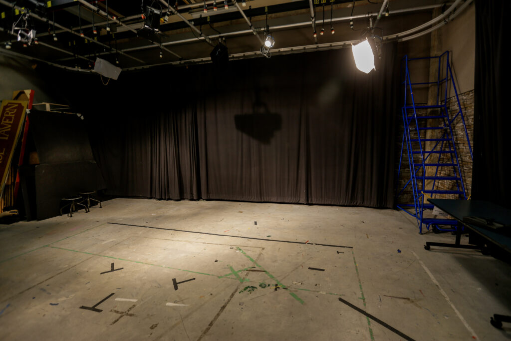 A working production studio featuring a concrete floor with stage blocking lines, dark brown curtains, overhead grid lighting, and scattered props like a blue rolling ladder and stacked set pieces.