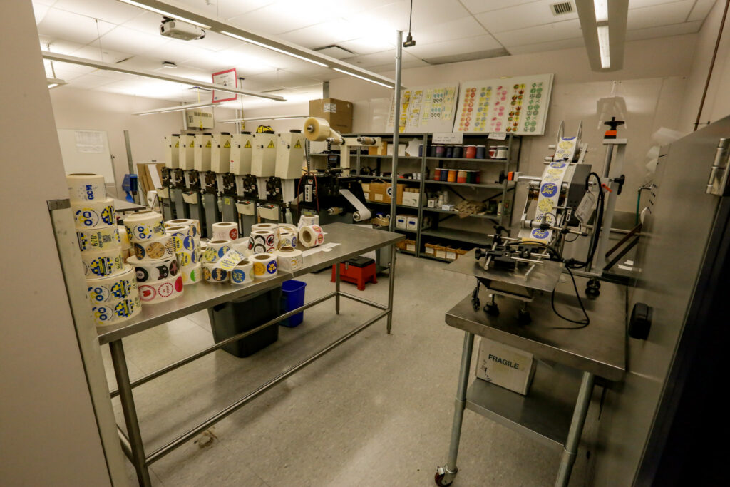 An industrial workshop setup for label processing, featuring a line of beige machines, label finishing equipment, and stacks of colorful label rolls on stainless steel workstations, with shelves of supplies in the background.
