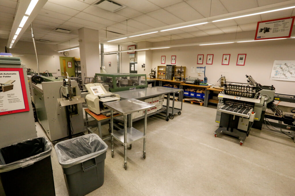 A well-lit print production lab featuring industrial paper folding, binding, and finishing equipment, with workbenches and informational posters.