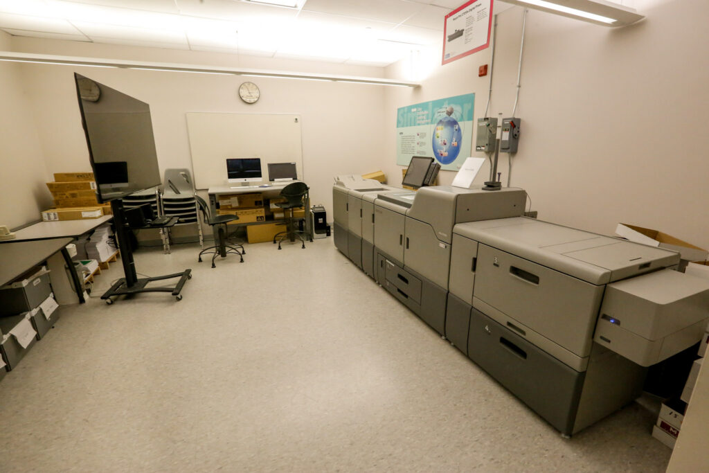 An brightly lit industrial print room featuring a large Ricoh Pro C7210x digital press on the right, and multiple computer workstations and storage on the left, used for printing services.