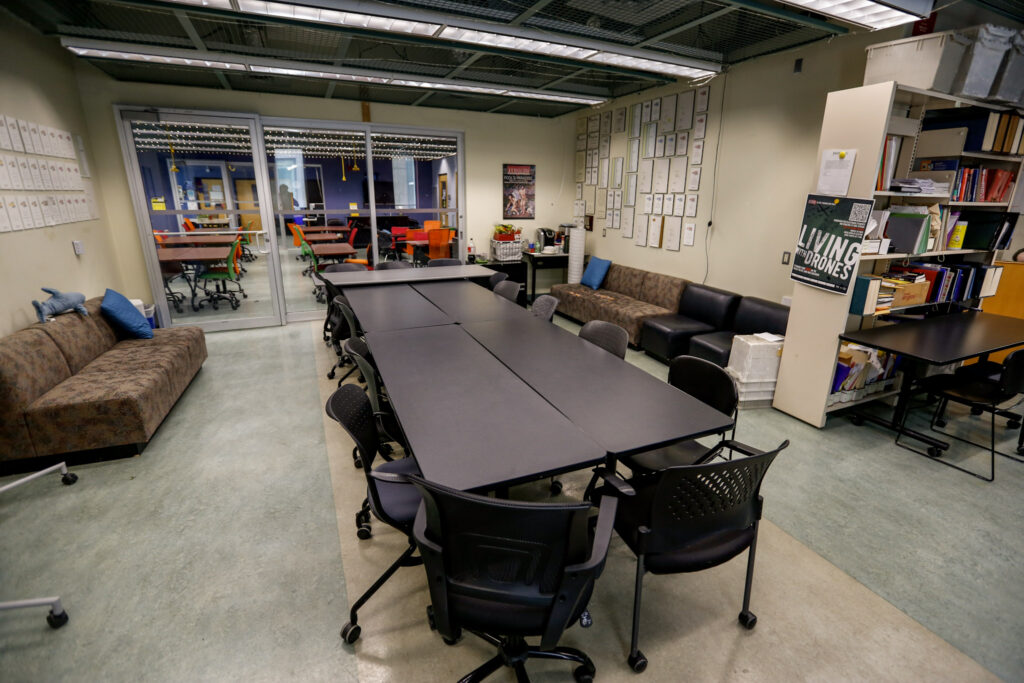 An academic or office common room featuring a large black meeting table, several couches for lounging, a coffee station, and bookshelves, with glass doors opening to another colorful workspace.