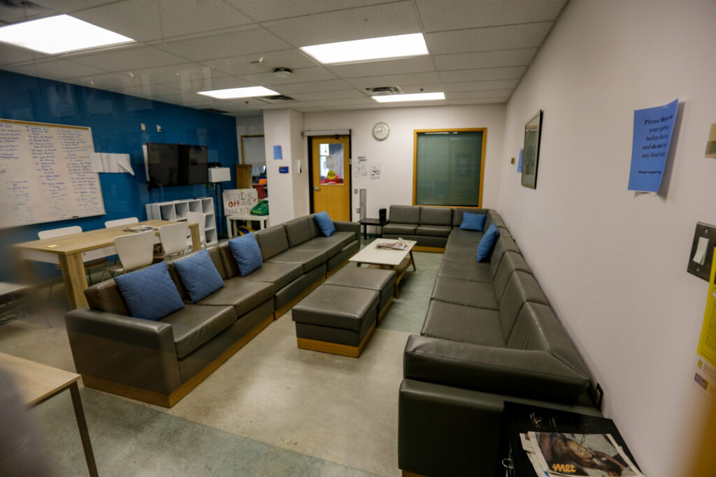 An interior shot of a spacious common room, featuring extensive dark grey modular sofas with blue throw pillows, a coffee table, and a dining area with a long wooden table and white chairs. One wall is bright blue with a whiteboard, while others are off-white.