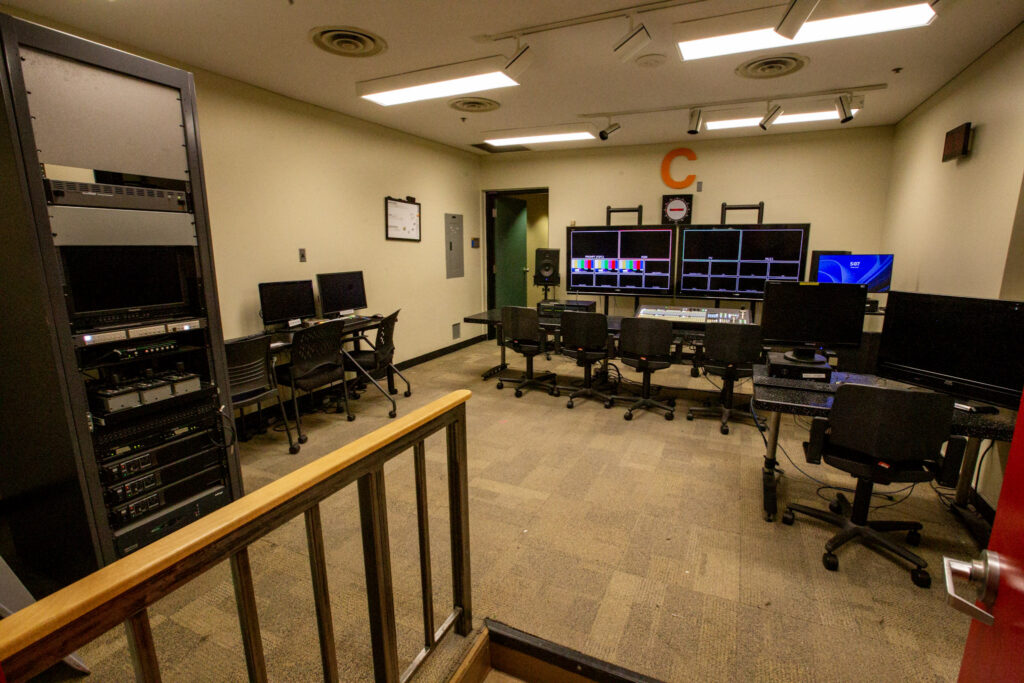 An unoccupied media production control room with multiple video monitors displaying feeds, an extensive equipment rack, and several operator workstations.