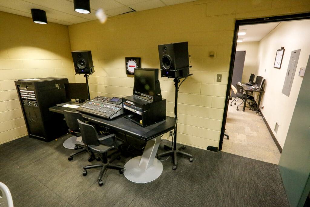 An audio production control room, featuring a large mixing console, monitors, and speakers, with a computer lab visible through an open doorway.