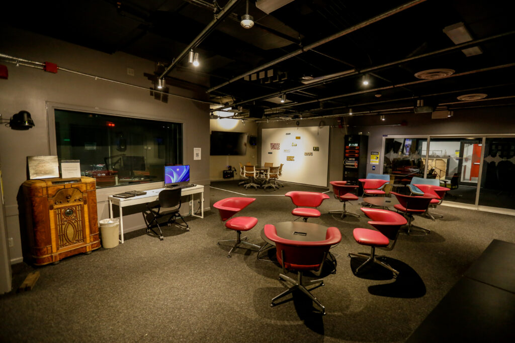 An eclectic, dimly lit media production and collaboration space featuring a historic radio console, a computer workstation, clusters of modern red swivel chairs, a conference table, and a soundproof studio visible through a large window. A whiteboard displays media-related terms.