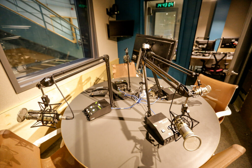 An unoccupied professional radio or podcast studio, featuring a round table equipped with four microphones on boom arms, individual control units, and a central monitor, ready for a multi-person broadcast.