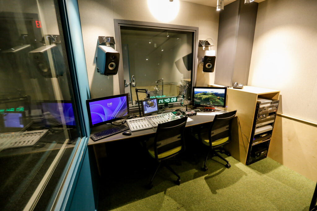 A professional audio studio control room featuring a desk with two mixing boards, multiple computer monitors, microphones, and wall-mounted speakers, with a soundproof window looking into an adjacent booth.
