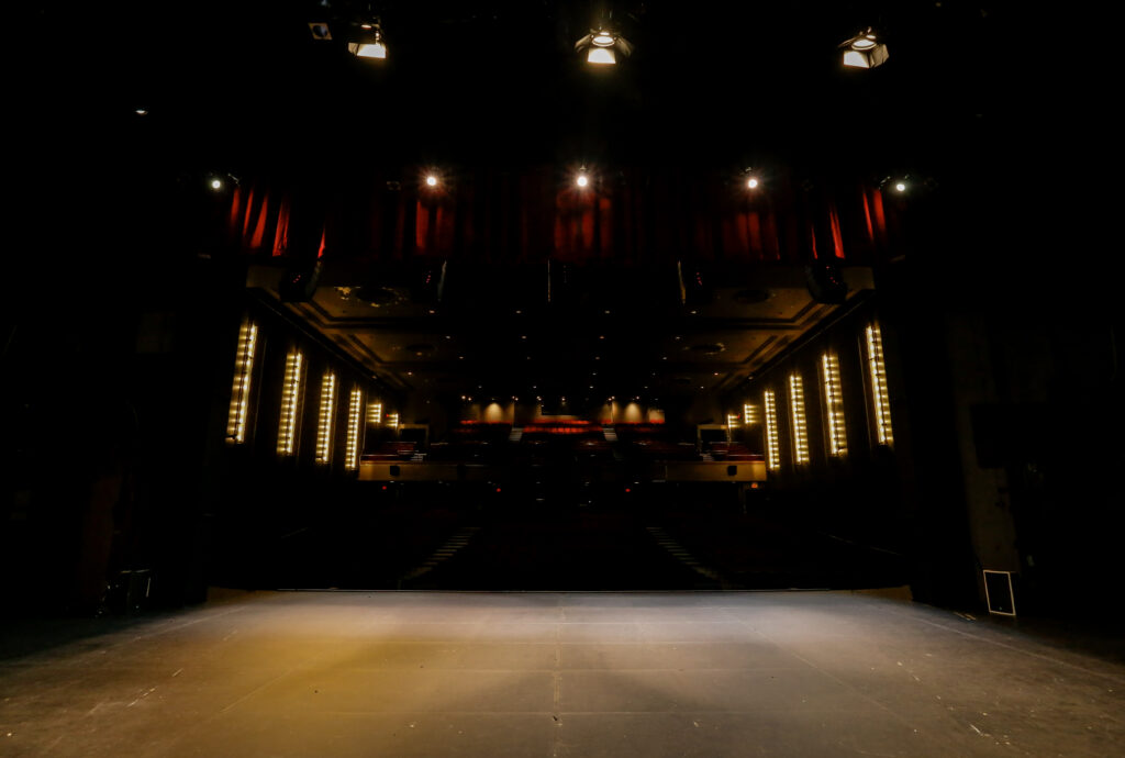 View from an illuminated stage towards a dark, empty theater auditorium with rows of red seats, framed by red curtains and illuminated by warm wall sconces.