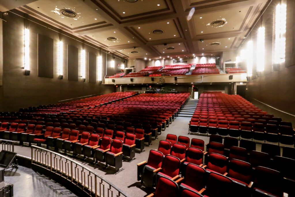 A wide shot of an empty theater auditorium featuring rows of red velvet seats, illuminated by tall vertical wall lights and overhead fixtures, with a balcony section visible in the background.