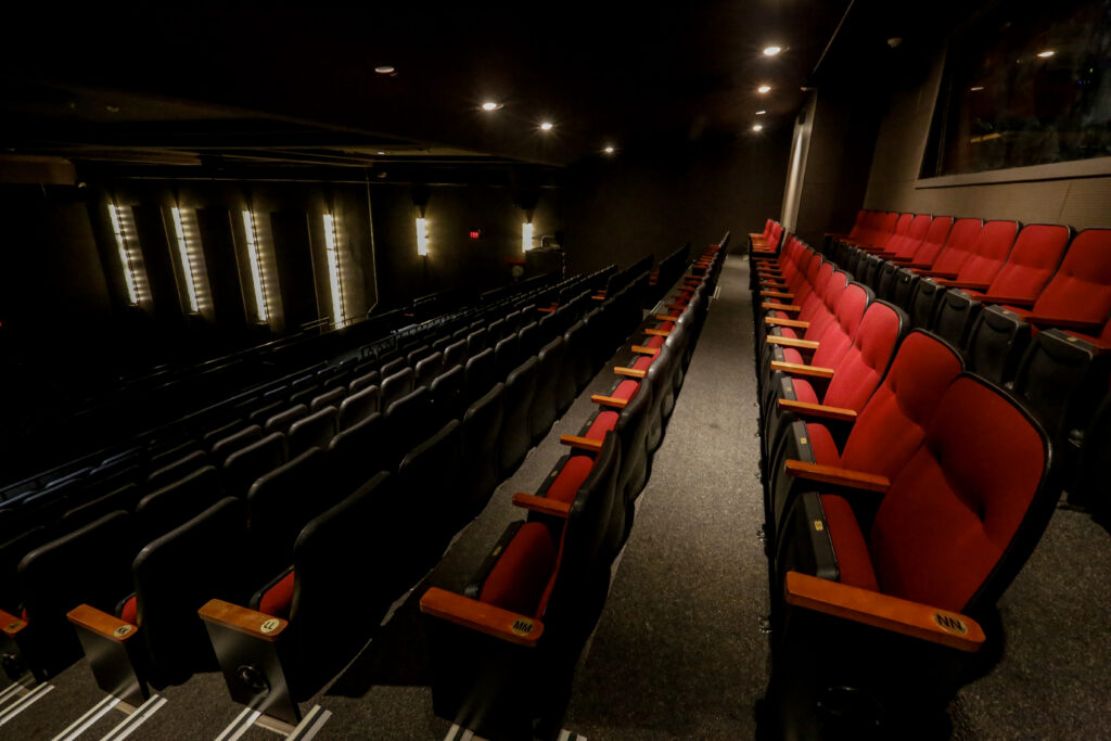 An empty theater or lecture hall with rows of red and dark seating, illuminated by overhead and wall lights, featuring a prominent exit sign.