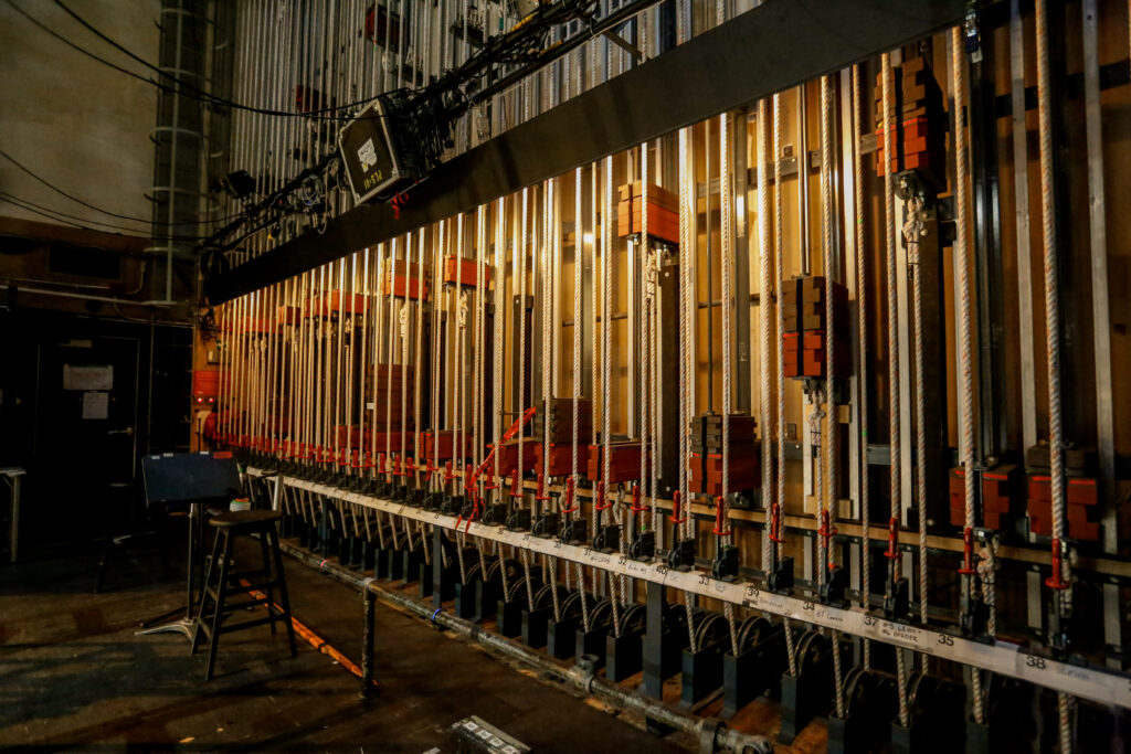 An interior shot of a theater's fly rail system, showing a long row of ropes with red-handled locks and counterweights used to raise and lower stage elements, with an operator's stool nearby.