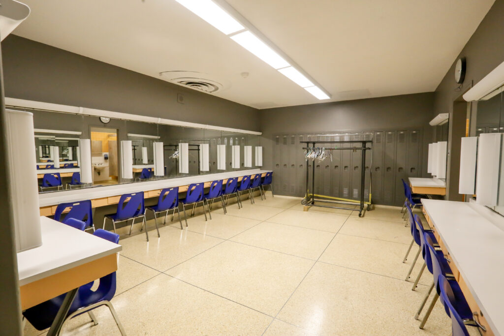 A spacious and brightly lit dressing room, likely for a theater or event, featuring long makeup counters with mirrors and blue chairs, alongside a wall of gray lockers and a mobile costume rack with hangers. A reflection shows an adjacent restroom.