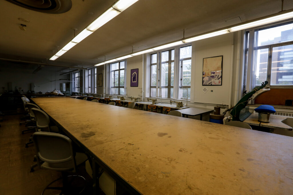 An empty fashion or textile workshop with a long central cutting table, a row of industrial sewing machines along large windows, and specialized pressing equipment.
