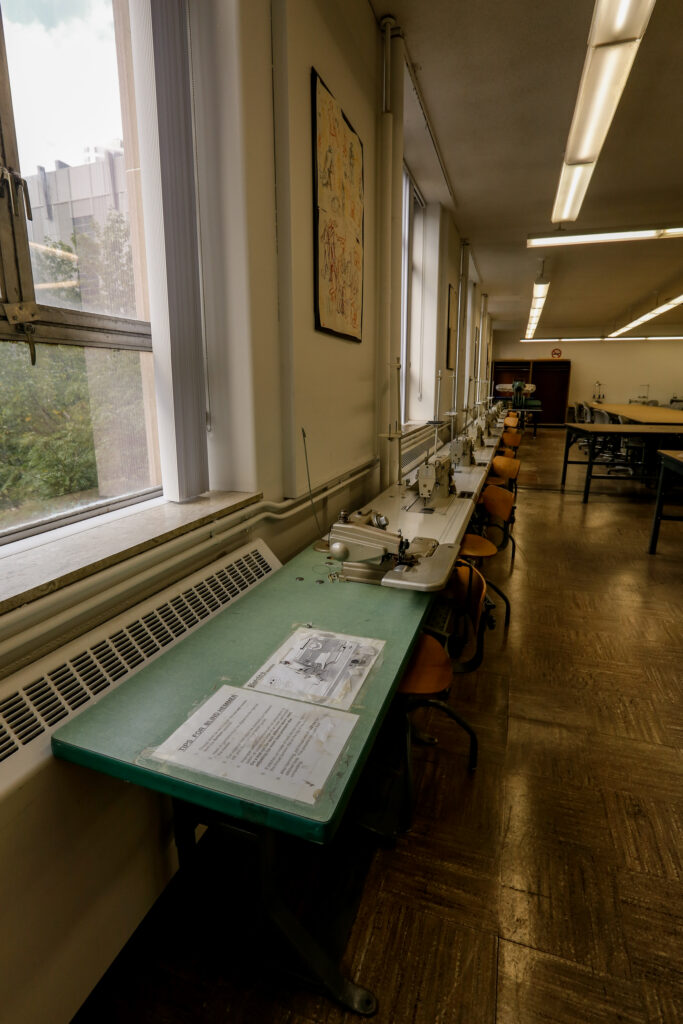 An industrial sewing lab featuring a long row of sewing machines on green tables, illuminated by natural light from large windows and overhead fluorescent lights.