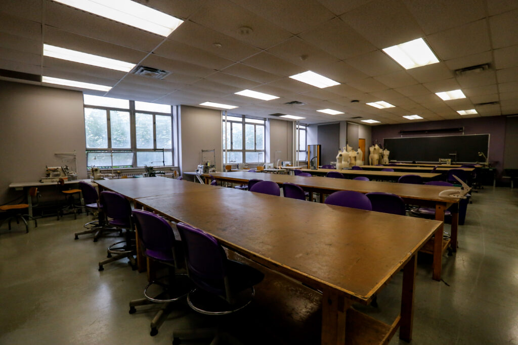 A fashion or textile design classroom with rows of long tables, purple chairs, industrial sewing machines by windows, and dress forms near a chalkboard.
