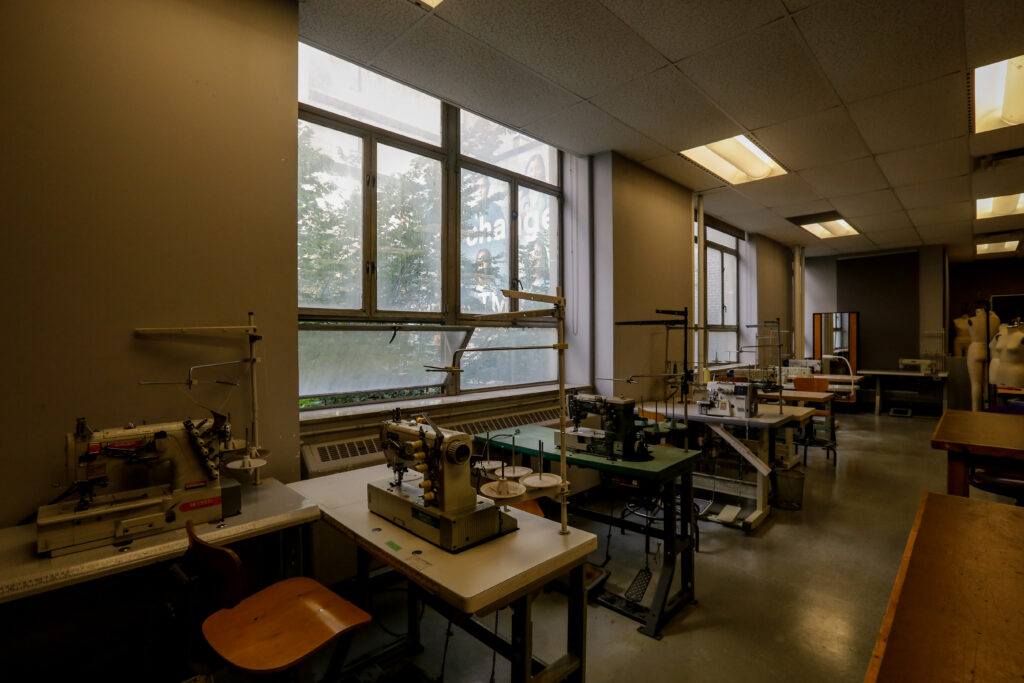 An organized fashion design or textiles classroom filled with various industrial sewing machines on tables, facing large windows, and dress forms in the background.