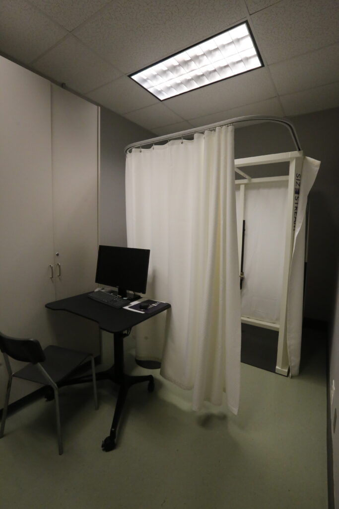 A room featuring a computer desk with a monitor and a chair, next to a private booth enclosed by white curtains, possibly a Size Stream body scanner or fitting area.