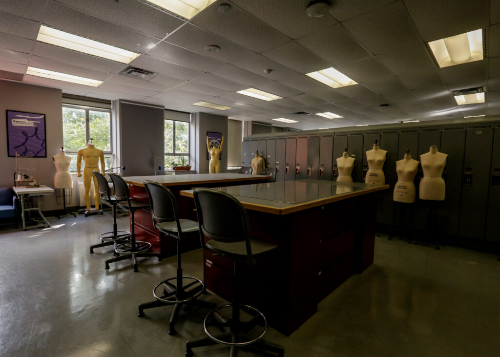 An empty fashion design classroom equipped with mannequins, work tables, a sewing station, and a wall of lockers, suggesting a creative learning environment.
