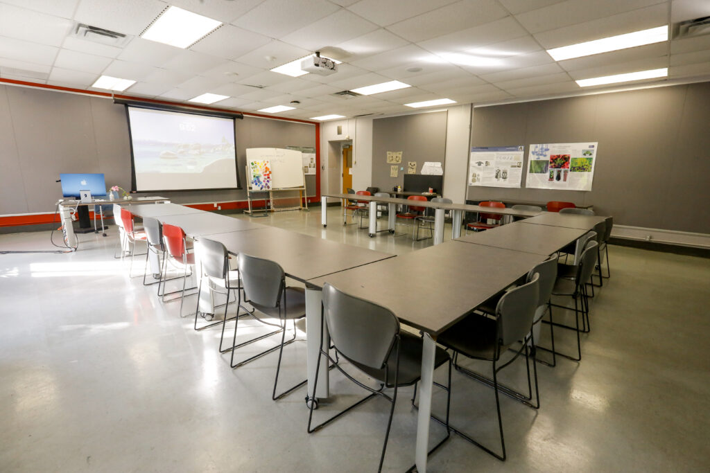 An empty, well-lit classroom or meeting room featuring modular tables arranged in a U-shape, ready for a presentation. It includes a projector screen displaying a landscape, a blue iMac, and various posters on the walls.