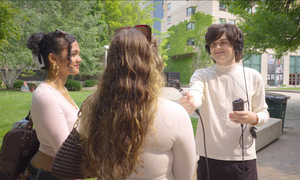 An outdoor interview in a sunny park or campus setting, with a young man holding a microphone towards a woman seen from behind, while another woman watches smiling.