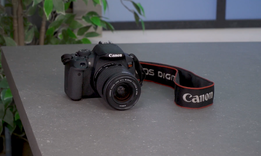 A black Canon EOS Rebel DSLR camera with an 18-55mm lens and its branded strap, resting on a speckled gray table.
