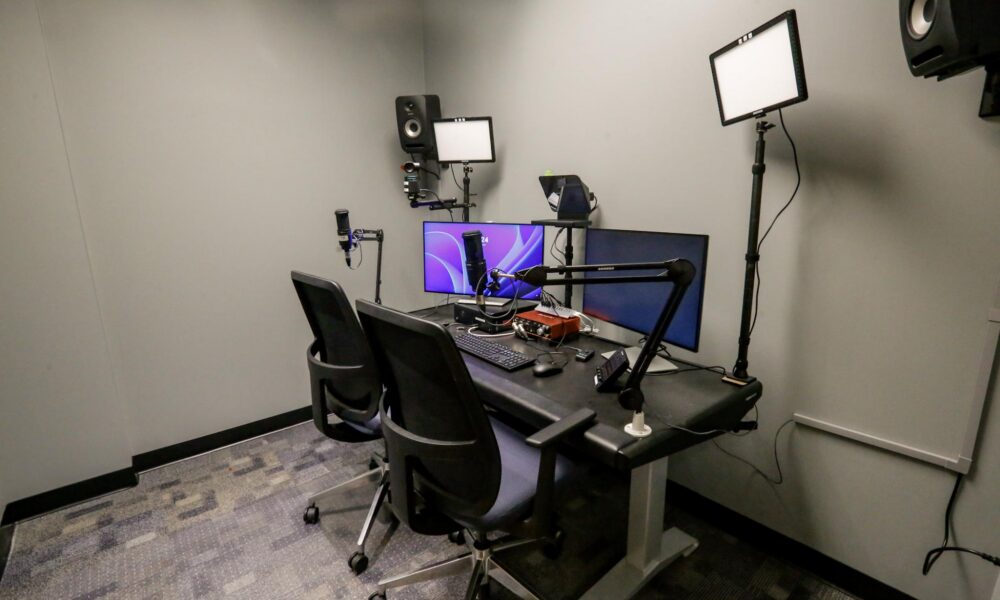 A modern podcast or streaming studio featuring a dual-person desk setup with monitors, professional microphones, an audio interface, and LED panel lights.